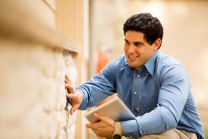 Latin descent building inspector, exterminator, architect, building contractor, engineer, or insurance adjuster examines a building/home's exterior stone wall. He holds a digital tablet.